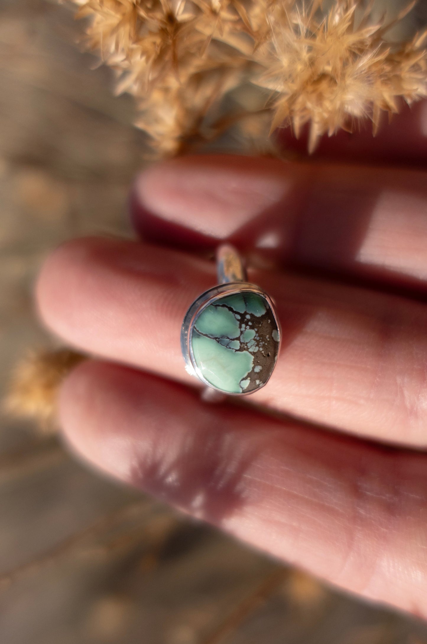 A hand holding a sterling silver ring with a green turquoise stone. The ring's shadow cast on the skin and there are dried florals in the soft background. 