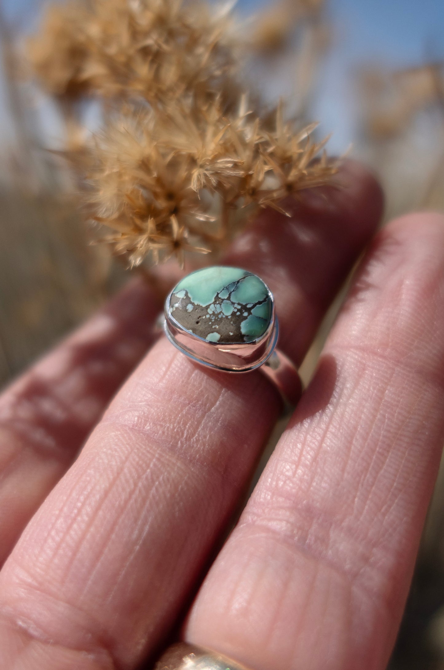 A handmade sterling silver and damele turquoise ring half by fingers outside in a field with dried florals and sky in the background