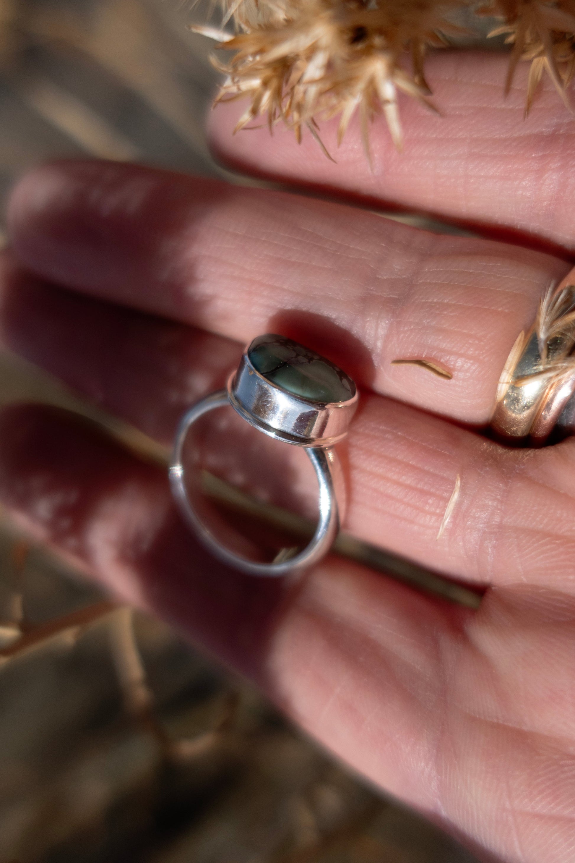Side view of a sterling silver ring from above half in a hand with an artistic scene of light and shadow with earthy tones and dried florals