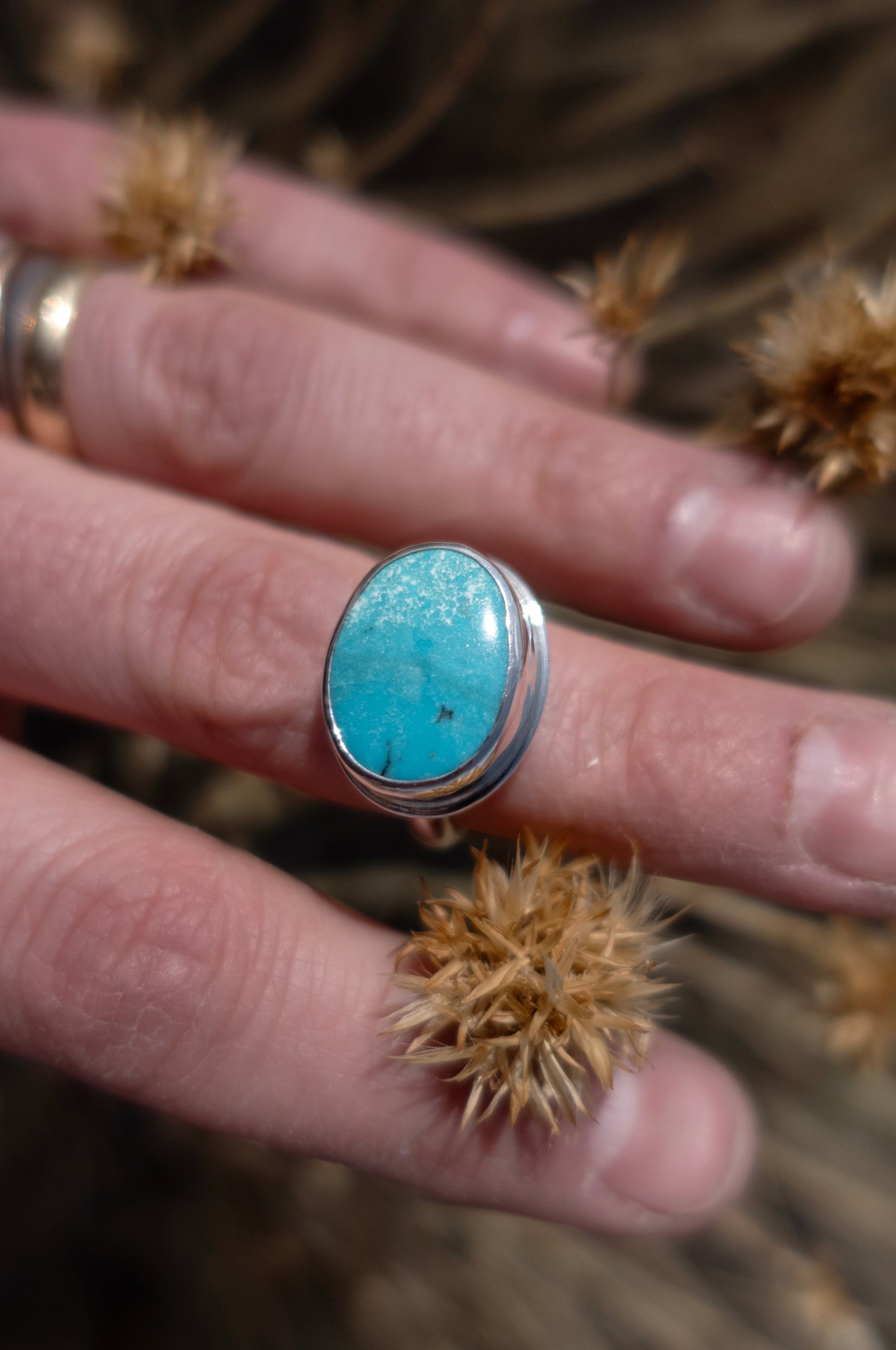A beautiful bright blue turquoise oil cabochon on a sterling silver handmade ring worn on a hand surrounded by pretty dried plants