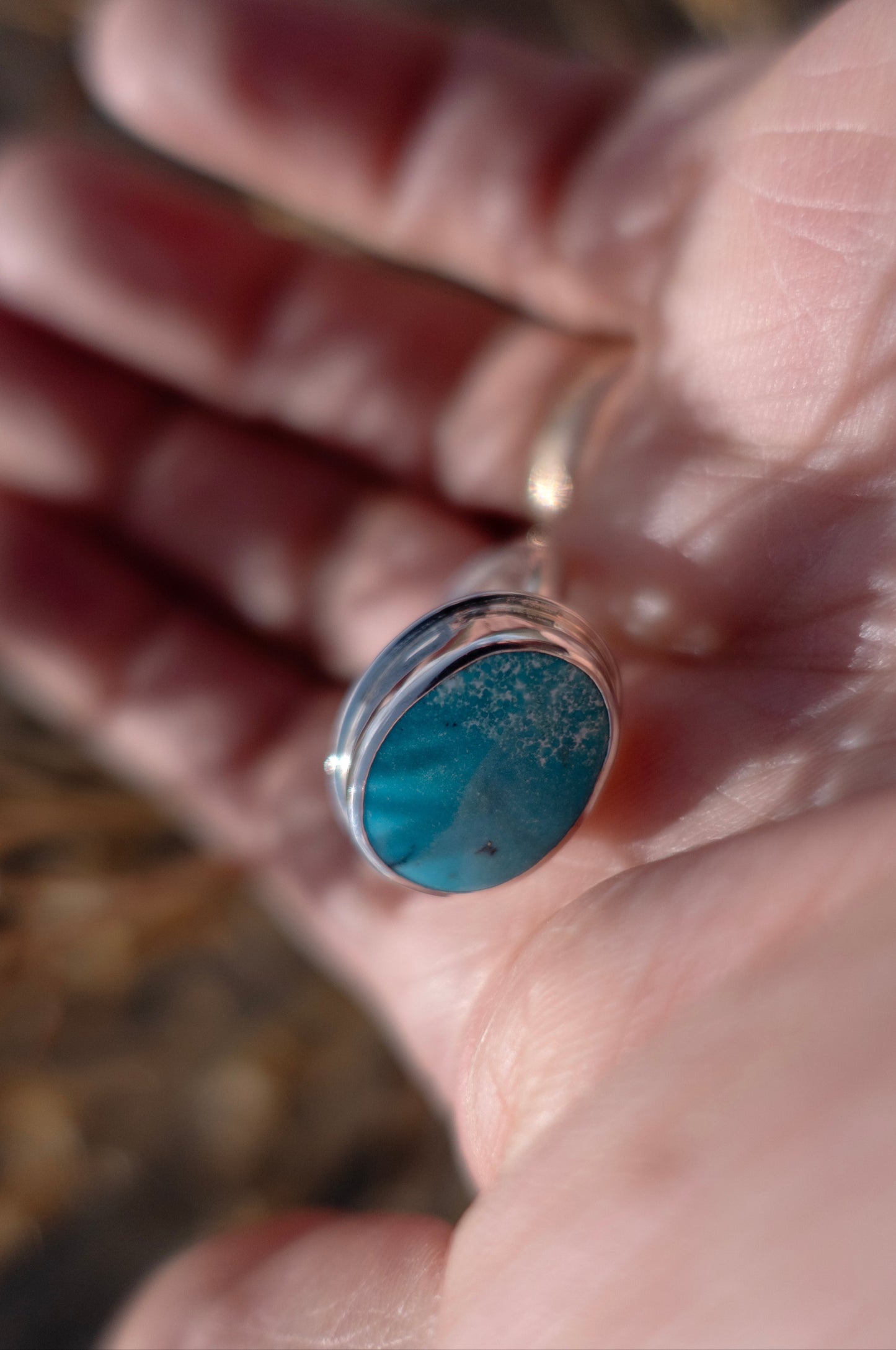 Delicate white speckles drift across this blue turquoise cabochon ring like a light dusting of snow. The handmade sterling silver ring is held in a hand with a soft background that is artistically out of focus.