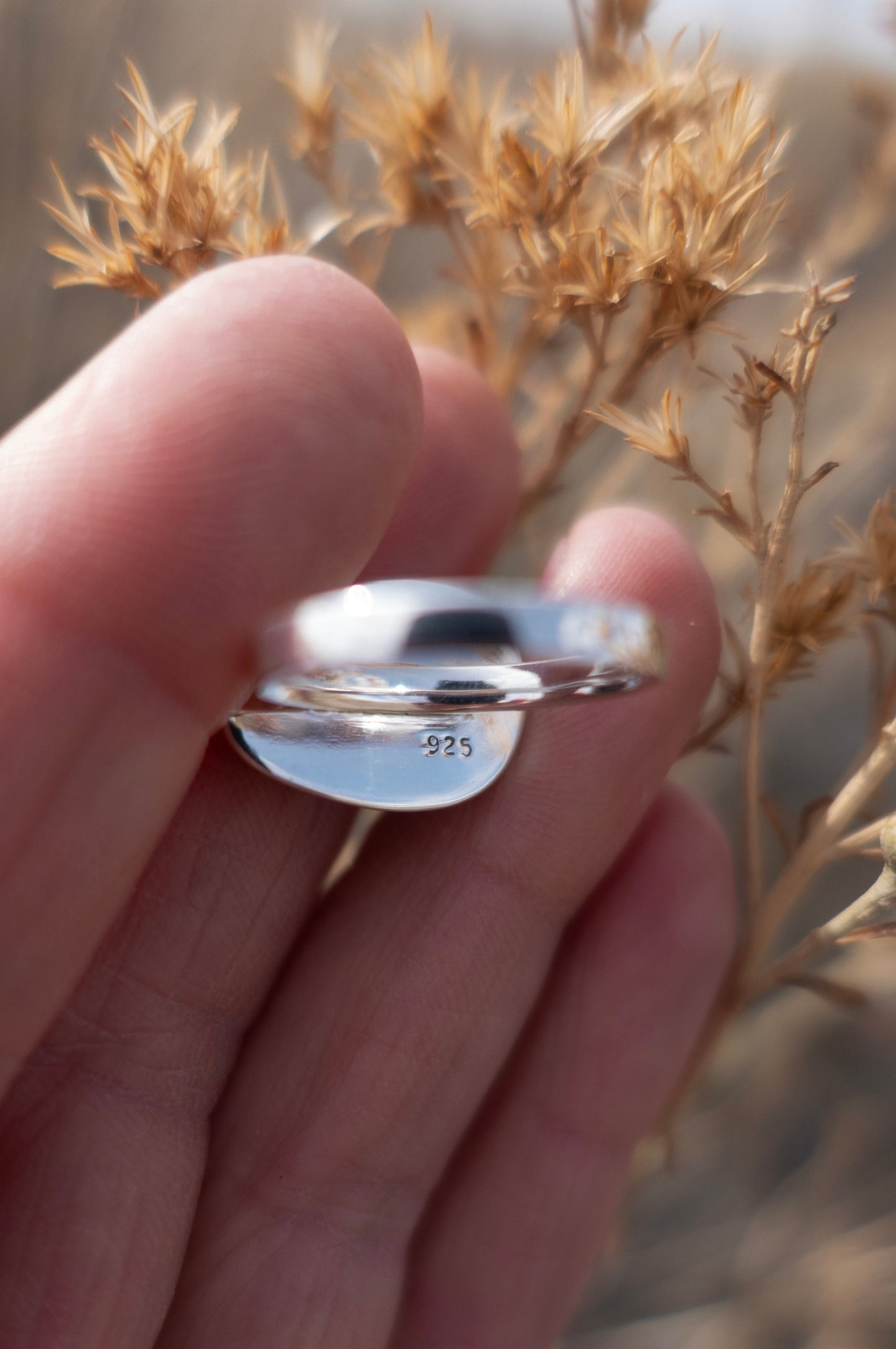 The inside of a delicate sterling silver ring that was made by hand and is stamped 925 held by fingers against an earthy floral background
