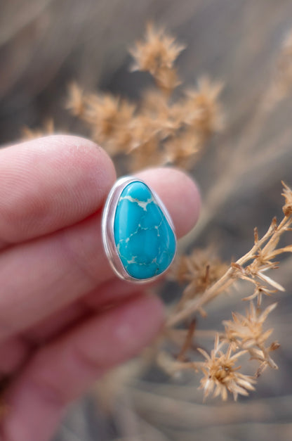 Glassy, vivid blue Kingman Turquoise cabochon and sterling silver ring that was made by hand. Held by hand and photographed in a soft focused earthy background with dried florals. 