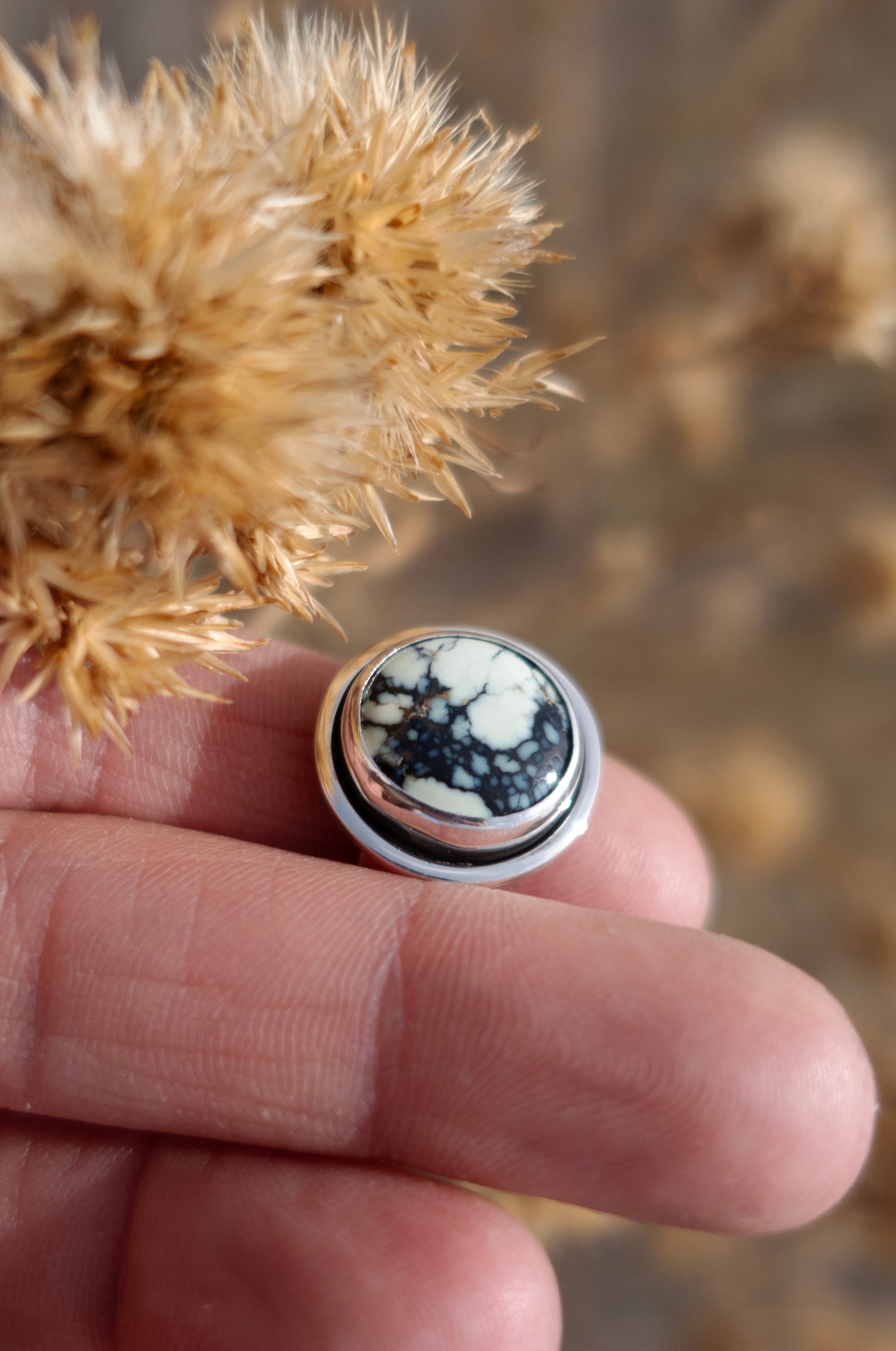 A finely polished sterling silver shadowbox style ring with a beautifully unique Poseidon Variscite cabochon on display. This ring is held by fingers and surrounded by dried plants and a earthy toned set background.