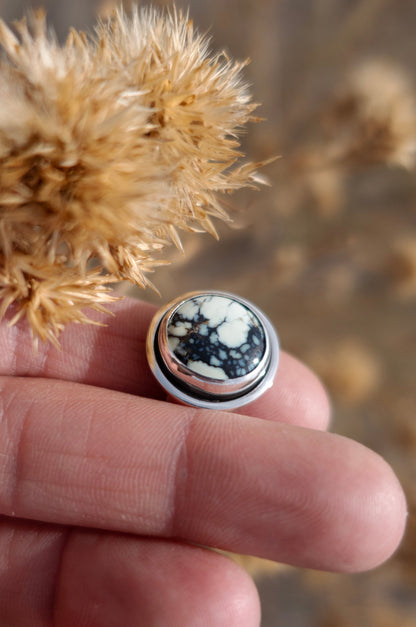 A finely polished sterling silver shadowbox style ring with a beautifully unique Poseidon Variscite cabochon on display. This ring is held by fingers and surrounded by dried plants and a earthy toned set background.