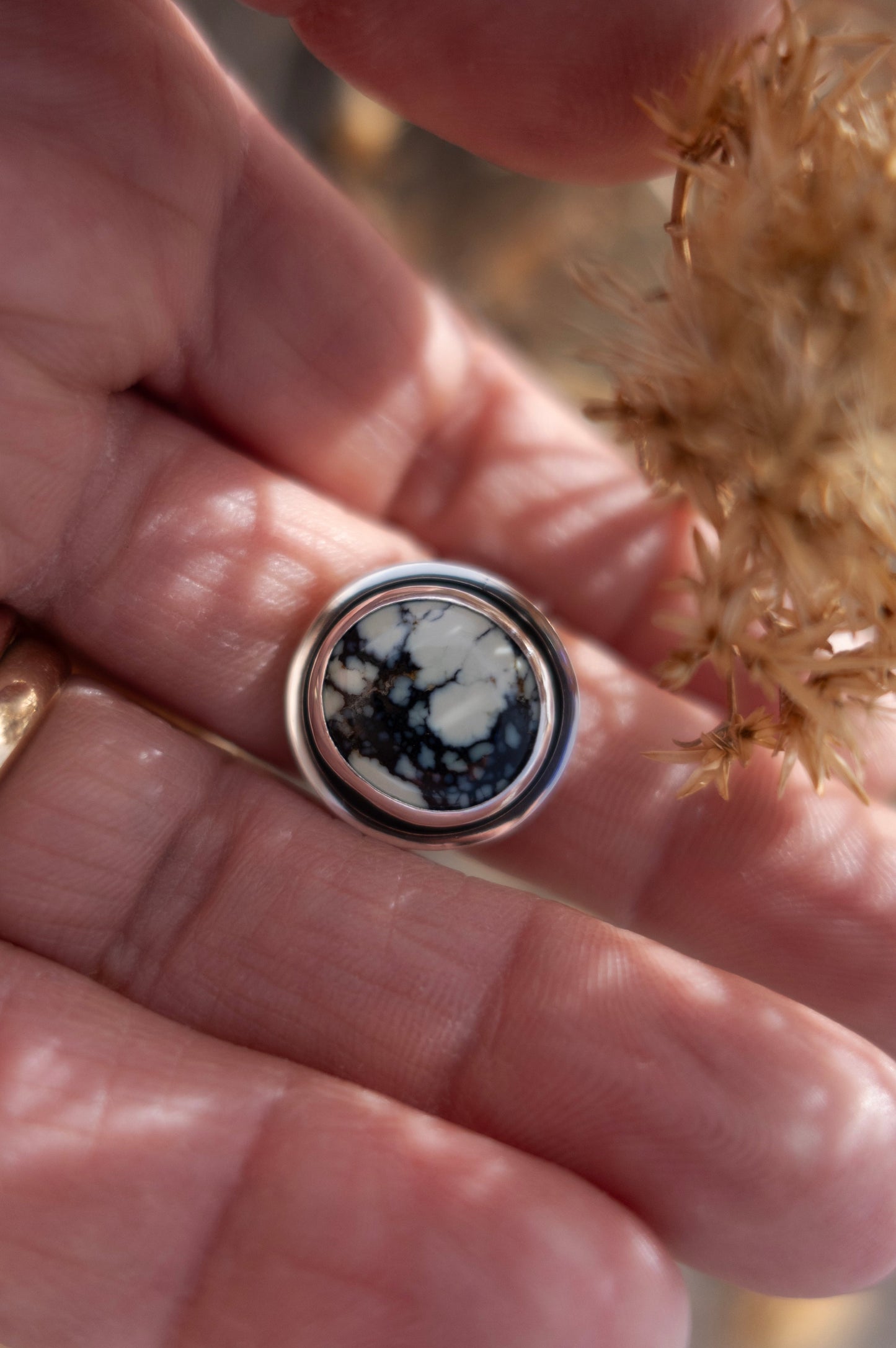 Close-up of a hand wearing a handmade sterling silver ring with a unique minty green Poseidon Variscite stone, beautifully surrounded by a shadowy soft background and dried plants.