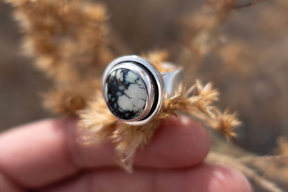Closeup detail of a handmade sterling silver ring with a shadowbox setting and a thick band. The stone is a glowing minty green color with pools of blue and green admits its natural webbed matrix. The ring is held up by fingers and is framed by soft earthy materials.