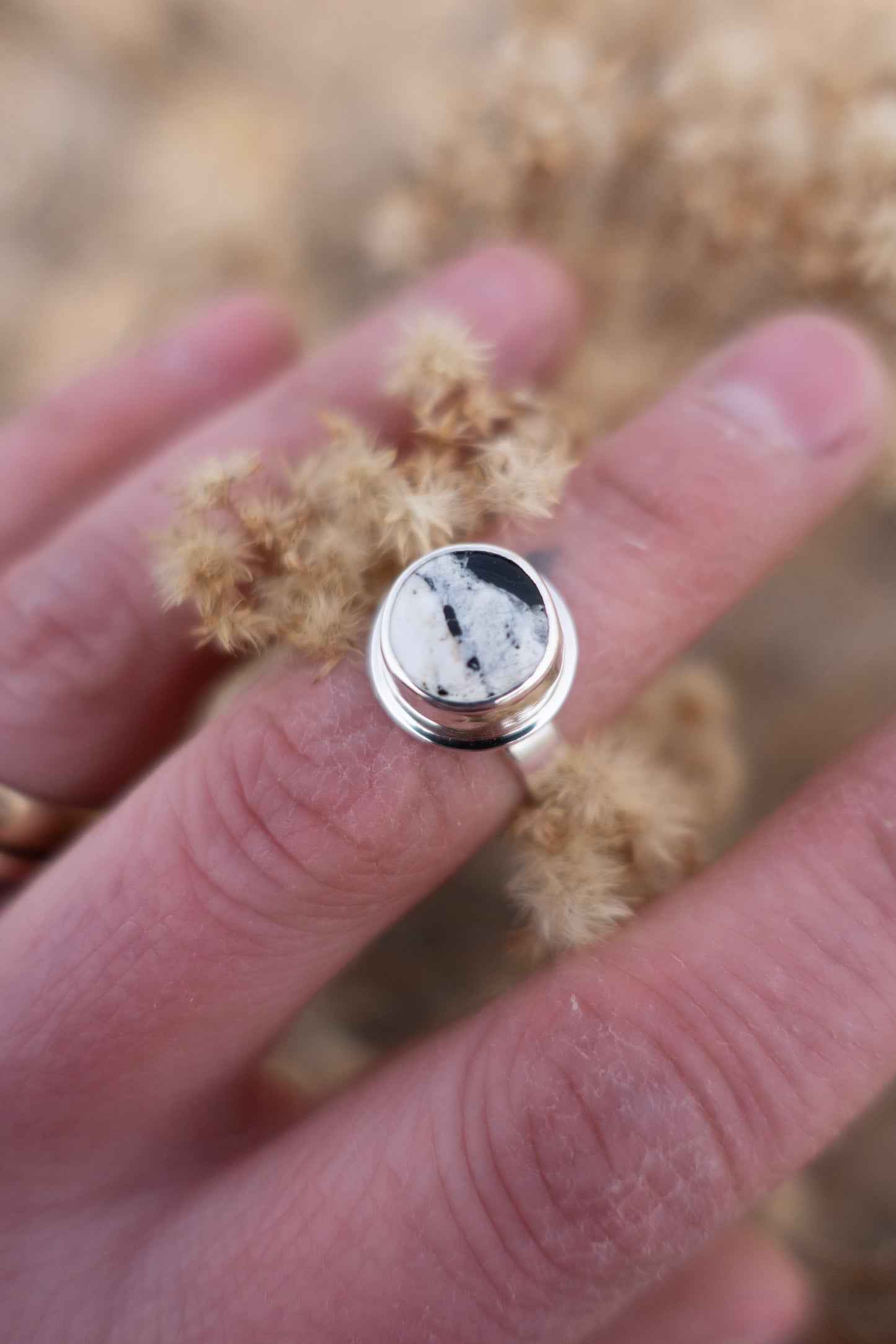 A silver ring with a round white buffalo turquoise is displayed on a person's finger, set against a backdrop of dried plants.