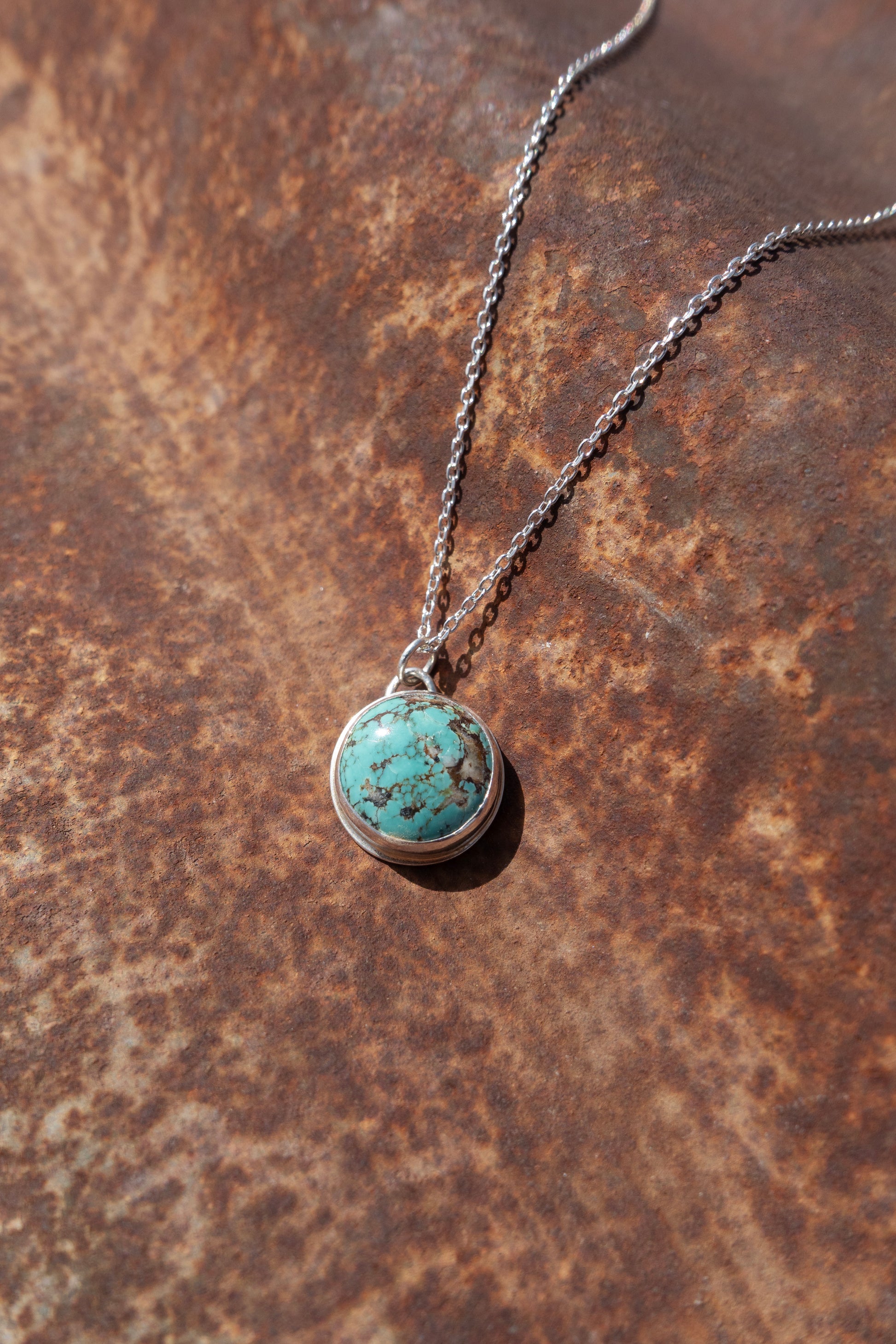 A round Kingman Turquoise handmade charm strung on a sterling silver cable chain displayed against a rustic background.