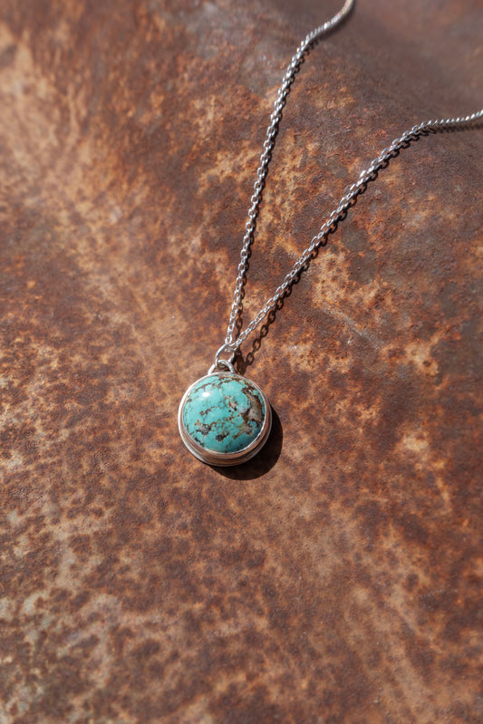 A round Kingman Turquoise handmade charm strung on a sterling silver cable chain displayed against a rustic background.