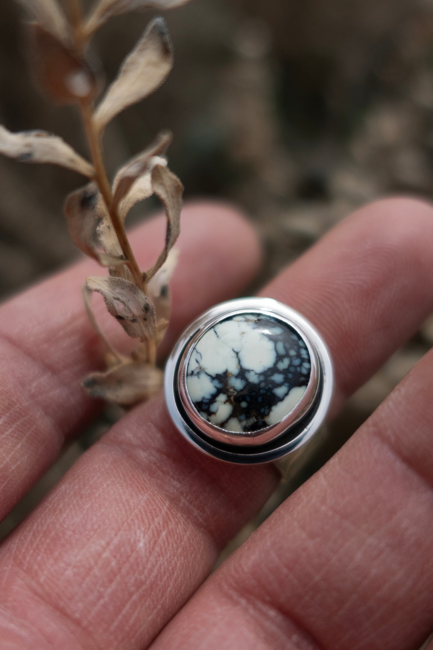 A sterling silver ring with a round Variscite stone in the center, held between fingers with a dry plant in the background.