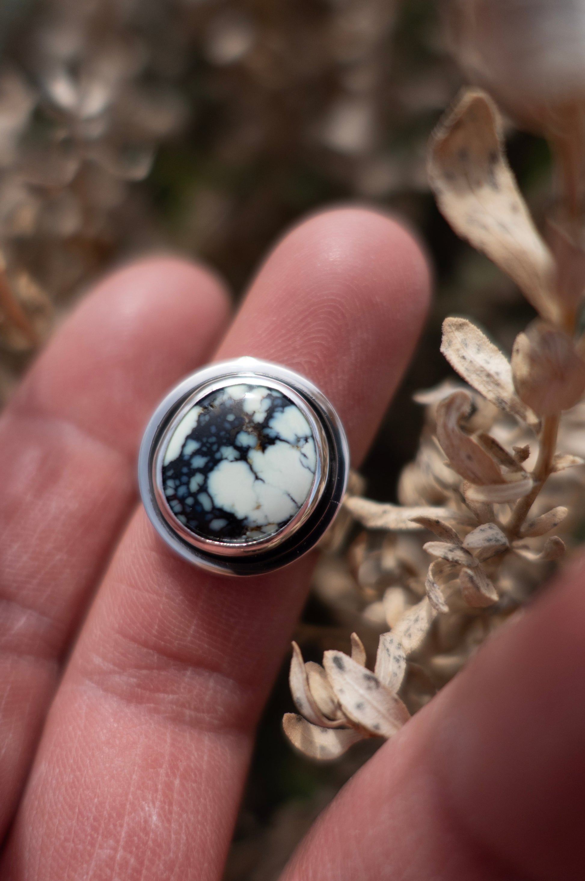 A beautiful shiny sterling silver ring handmade with skill and care. The circular design showcases a unique minty green and blue Poseidon variscite cabochon framed by a shadowbox style setting. This stunning ring is held by fingers amidst a soft earthy background.