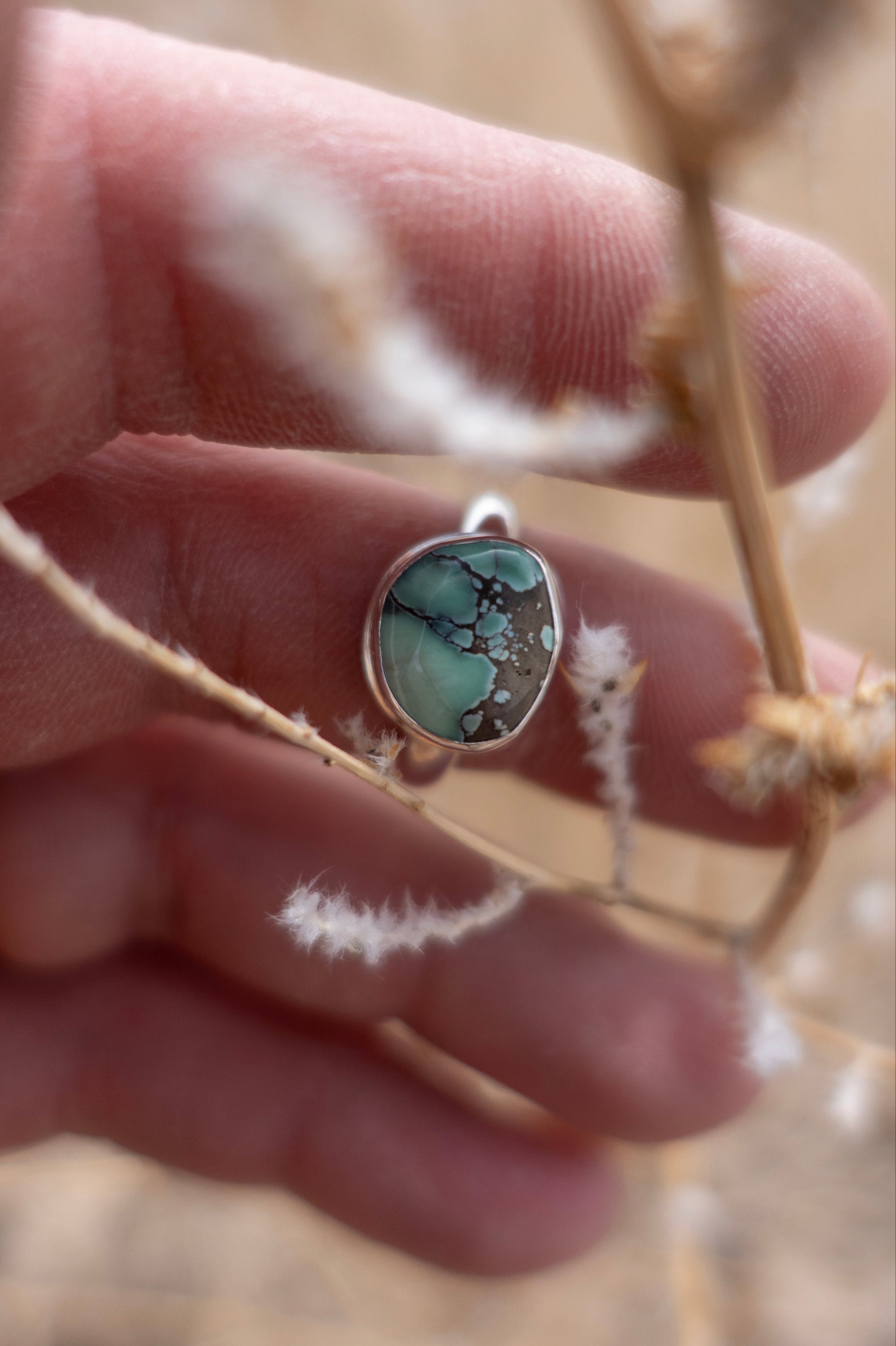 A hand holding a handmade sterling silver ring with a turquoise stone set in a half round design, displayed against a natural background with dried plants.
