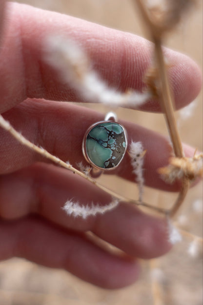 A hand holding a handmade sterling silver ring with a turquoise stone set in a half round design, displayed against a natural background with dried plants.